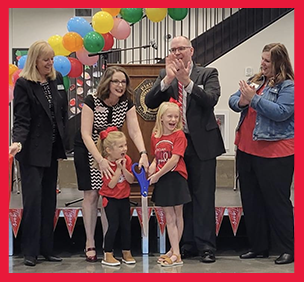 Ribbon cutting ceremony with clapping adults, two little girls, large scissors, and balloons in the background